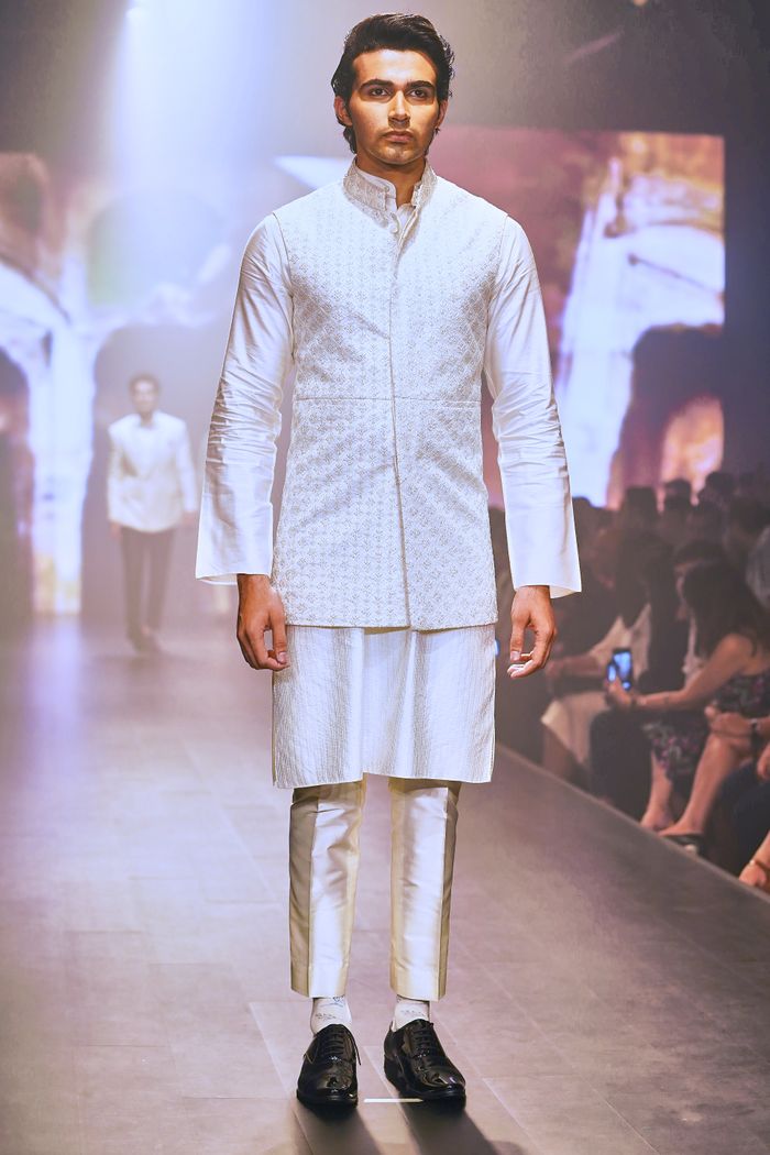 Male model in white traditional attire walking a runway during a fashion show.
